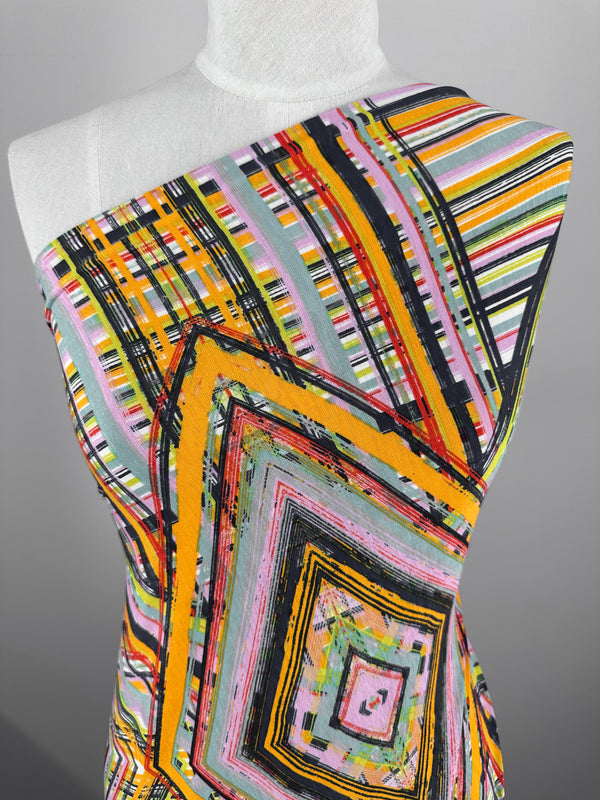 The mannequin displays the Super Cheap Fabrics' Printed Lycra - Geo Dome, featuring colorful geometric patterns of stripes and squares in orange, pink, black, yellow, and green. It's presented at an angle in soft lighting against a neutral backdrop. Width: 145cm.