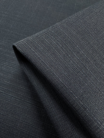 A neatly folded close-up of Super Cheap Fabrics' Textured Bengaline in Textured Indigo reveals its dark gray, medium-weight stretch with subtle horizontal lines and indigo threads. The fine weave and slight sheen contribute to a modern, sophisticated look. Width: 145cm.