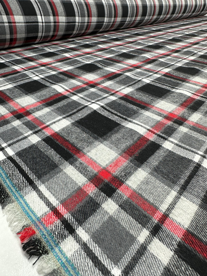 Displayed is a roll of Suiting - Tartanic - 147cm from Super Cheap Fabrics, featuring a plaid pattern with black, white, gray, and red intersecting stripes. The versatile fabric has a textured appearance and is spread out on a flat surface, with the roll partially unrolled in the background.