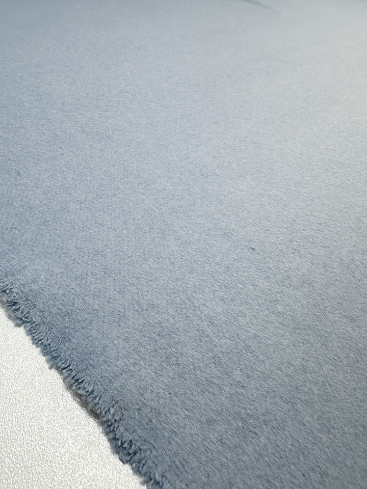 A close-up of the Wool Cashmere - Niagara Mist from Super Cheap Fabrics reveals a soft, light blue texture. The slightly frayed edge contrasts subtly with the smooth surface, while lighting accentuates its cozy and plush appearance, exuding luxurious softness.