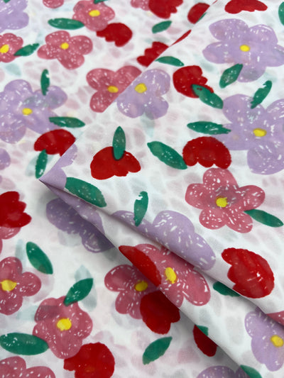 A close-up of Super Cheap Fabrics' "Salt Shrunk Print - Daise - 155cm" features a vibrant floral pattern with red, pink, and purple flowers amid lush green leaves on a white background. The salt shrunk treatment adds texture as the fabric gently folds.