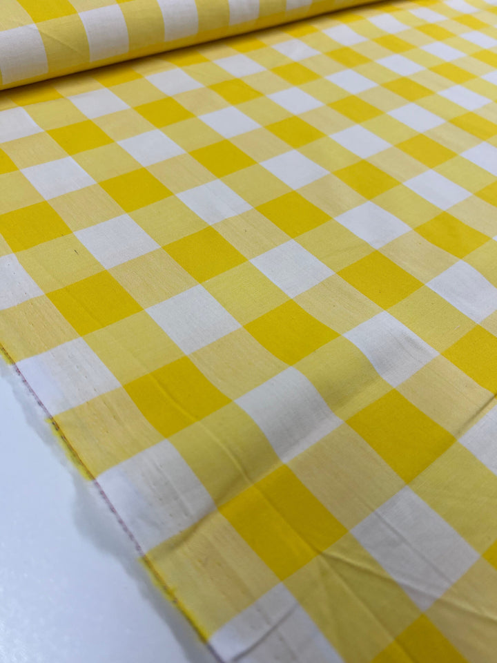 A detailed view of Cotton Gingham XL - Yellow - 112cm by Super Cheap Fabrics, showcasing a classic yellow and white gingham pattern made from breathable cotton fibers. The fabric is rolled out on a surface, emphasizing its smooth texture and vibrant colors.