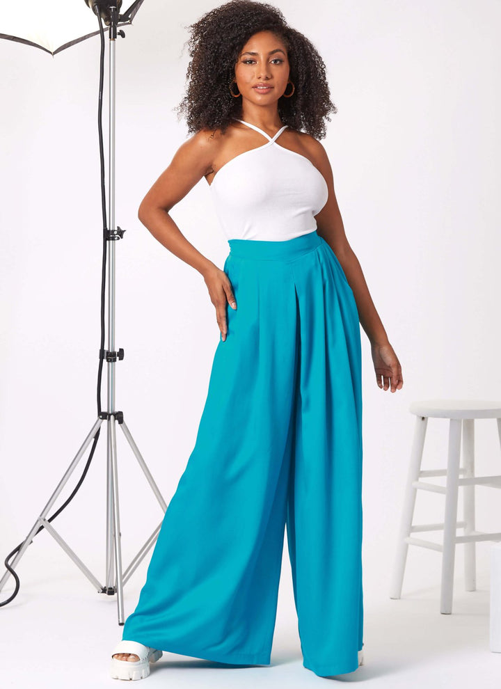 In a studio setting, a woman elegantly poses in pristine white fabrics and flowy turquoise trousers crafted using the New Look 6756 pattern for Misses’ Shorts and Trousers by Super Cheap Fabrics. She stands near a white stool and a lighting setup, enveloped by a soft, neutral background.