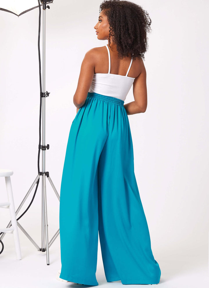 A woman with curly hair faces away, highlighting her chic halter-neck draped top and flowing pull-on trousers in a vibrant turquoise hue. She stands near a stool and studio light set against a white background, effortlessly exuding elegance in her stylish ensemble, captured beautifully by Super Cheap Fabrics' Pattern - New Look - 6756 - Misses’ Shorts and Trousers.