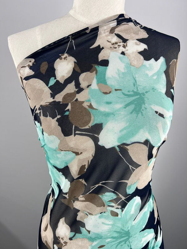 A mannequin is draped with Super Cheap Fabrics' Printed Mesh - Lilly, a lightweight fabric showcasing a bold floral pattern in shades of turquoise, tan, and white on a black background. The sheer 145cm wide fabric is wrapped asymmetrically over one shoulder, highlighting the large, intricate flower designs.