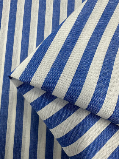 A close-up view of blue and white striped fabric. The 100% cotton cloth is folded, showing the vertical stripes pattern, which alternates between blue and white. The texture and weave of this lightweight fabric are visible, giving a sense of its material quality ideal for clothing. This is the Cotton Lawn - Blue Stripe - 150cm by Super Cheap Fabrics.