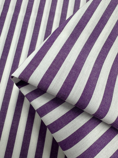 Close-up of a purple and white striped Cotton Lawn - Purple Stripe - 150cm fabric by Super Cheap Fabrics, featuring alternating vertical stripes. Part of the lightweight fabric is folded over, showcasing the pattern from different angles. The texture and weave of the 100% cotton material are visible, with the colors appearing vibrant.