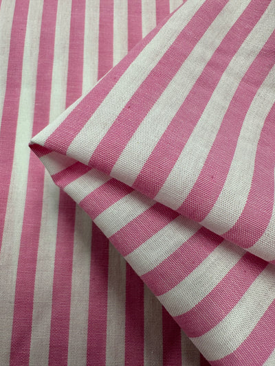 Close-up of a piece of lightweight fabric with vertical pink and white stripes. The 100% cotton material is slightly folded, showcasing multiple layers and the texture of the fabric. This is Cotton Lawn - Pink Stripe - 150cm by Super Cheap Fabrics.