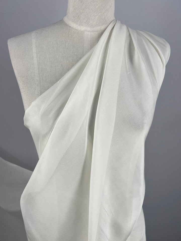 A white dress form is draped with a sheer, single-shoulder fabric made from Super Cheap Fabrics' elegant, extra lightweight Pure Silk Chiffon in platinum. The material is pleated and flows diagonally across the form, showcasing smooth, clean lines. The background is a plain, neutral grey, putting full emphasis on the luxurious 140cm silk fabric.