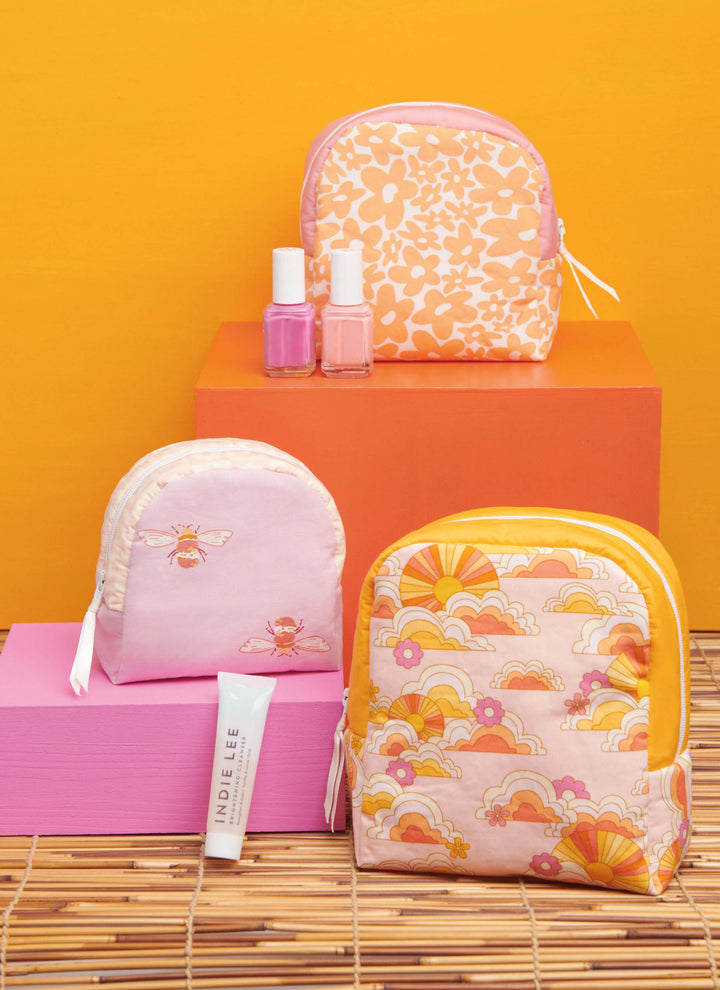 Displayed against a vibrant orange-themed backdrop are a set of three uniquely sized floral and patterned zipper cases from Super Cheap Fabrics' Pattern - Simplicity - 9937 collection. Accompanying these stylish accessories are two nail polish bottles and a skincare product tube, artfully arranged on stacked blocks, capturing the playful elegance reminiscent of empire waist tops.