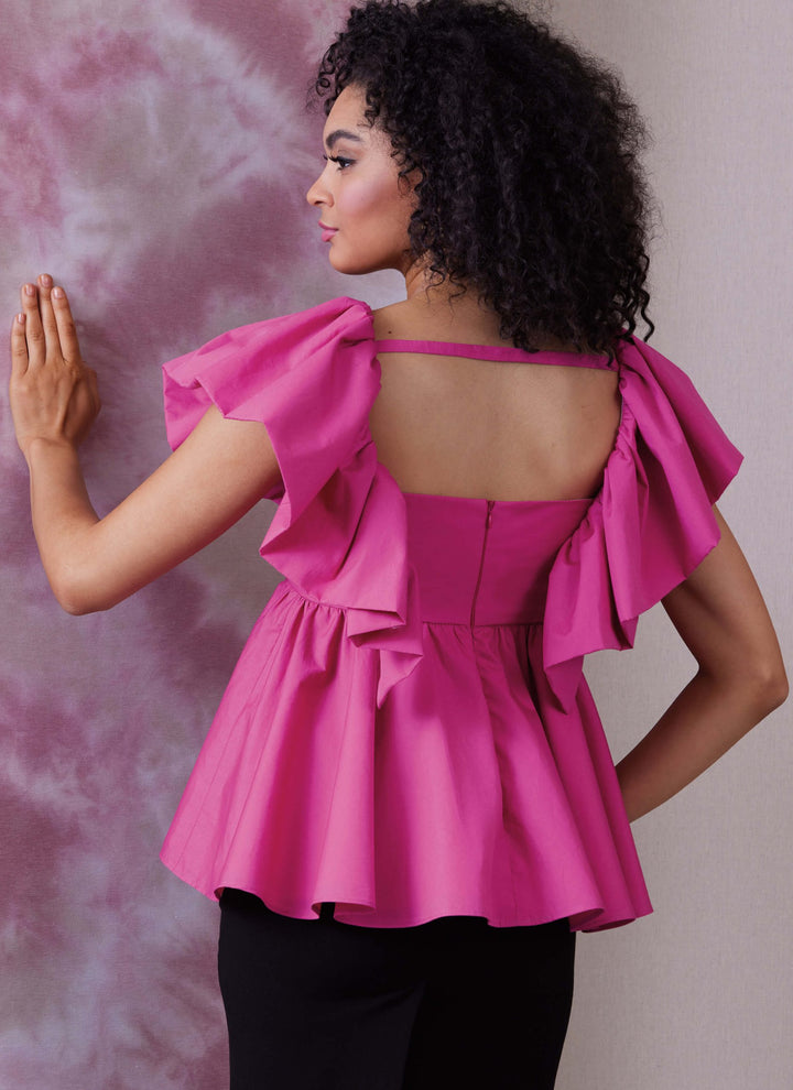 A woman with curly hair stands against a textured purple and gray background, wearing a vibrant pink, ruffled sleeveless top from the Pattern - Vouge - 1940 collection by Super Cheap Fabrics, paired with black pants. Her left hand rests on the wall as she gazes to her left.