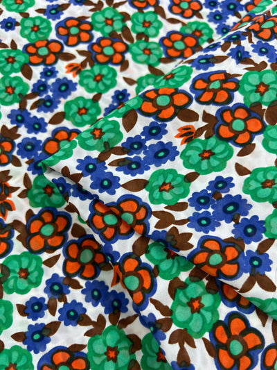 A close-up of the Salt Shrunk Print - Kaleid from Super Cheap Fabrics shows an elegant, vibrant floral pattern with large green and brown flowers, smaller blue flowers, and orange accents on a white background. The fabric's folds highlight its intricate, vivid design. Width: 155cm.