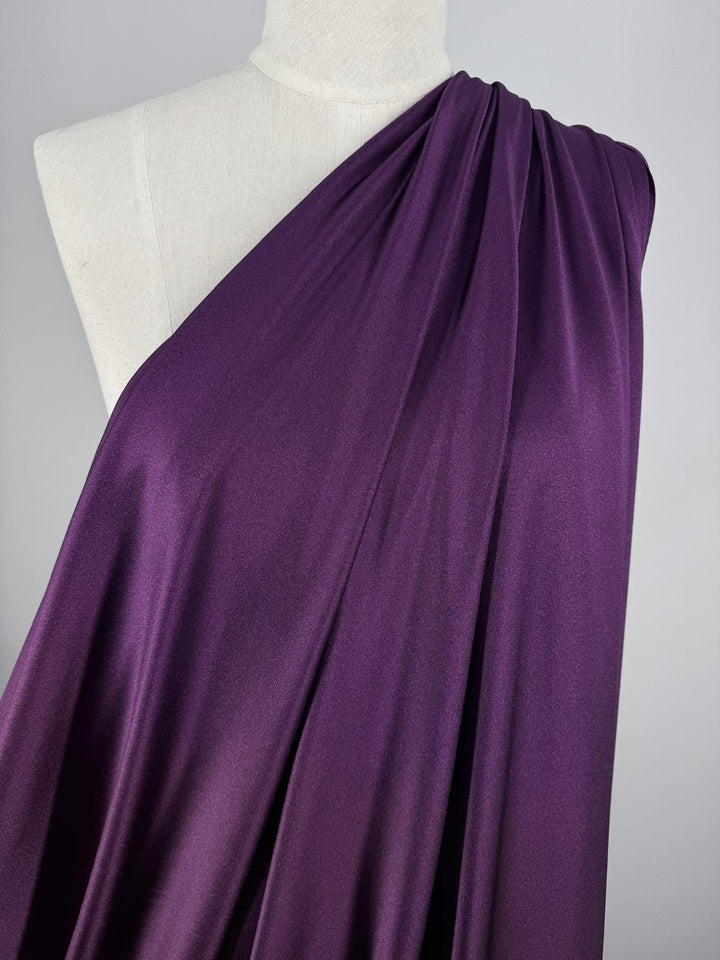 A white mannequin showcases Super Cheap Fabrics' Nylon Lycra - Winter Bloom (154cm), a smooth, deep purple fabric elegantly draped over one shoulder against a plain light gray background.