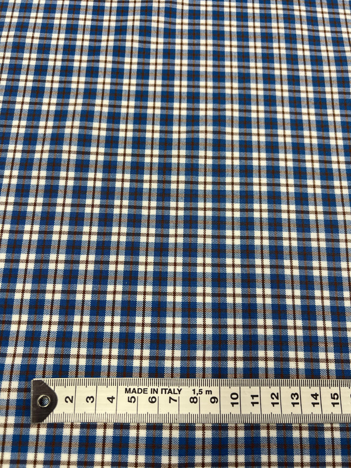 A close-up of the Shirting - Ralph - 150cm from Super Cheap Fabrics, featuring blue, white, and black plaid polyester fabric, highlights its versatile applications. A measuring tape at the bottom indicates a length of 18 centimeters.