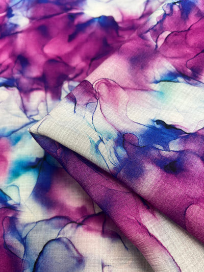 A close-up of the Designer Cotton - Celeste by Super Cheap Fabrics shows a luxurious, soft cotton material with an abstract, watercolor-like pattern in hues of purple, pink, blue, and white. The 150cm wide fabric is neatly folded and slightly translucent—perfect for luxury sewing projects.