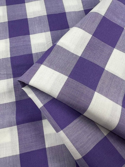 A close-up view of the Cotton Gingham XL - Sand Verbena from Super Cheap Fabrics captures the folded, neatly arranged pattern of large purple and white squares in this classic printed cotton fabric. The natural cotton fibers highlight a simple design that lends itself to versatile uses.