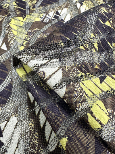 A close-up of Super Cheap Fabrics' Designer Mesh - Direction, a 150cm folded polyester fabric with a geometric pattern in yellow, brown, and black. Ideal for home décor, it features detailed texture with visible stitching and intricate designs.