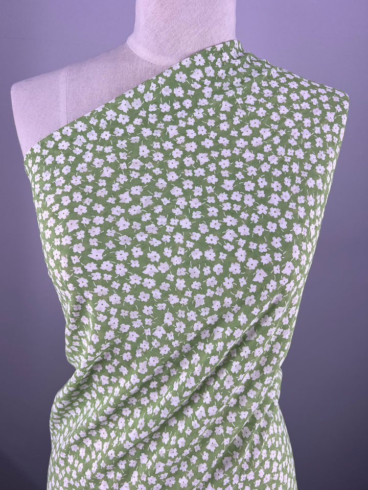 A dress form elegantly draped with Super Cheap Fabrics' Salt Shrunk Print - Jassy (155cm), featuring a pattern of small white flowers on green fabric, arranged asymmetrically against a purple backdrop to highlight its vibrant prints.