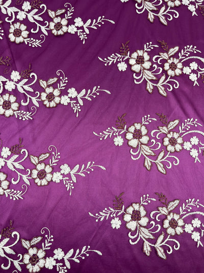 A purple fabric with embroidered floral patterns in white and beige. The intricate designs are evenly spaced, featuring flowers, leaves, and swirling vines. This Super Cheap Fabrics Embroidered Lace - Burgundy - 150cm lightweight fabric adds a delicate and elegant touch to the material, reminiscent of burgundy lace arrangements.