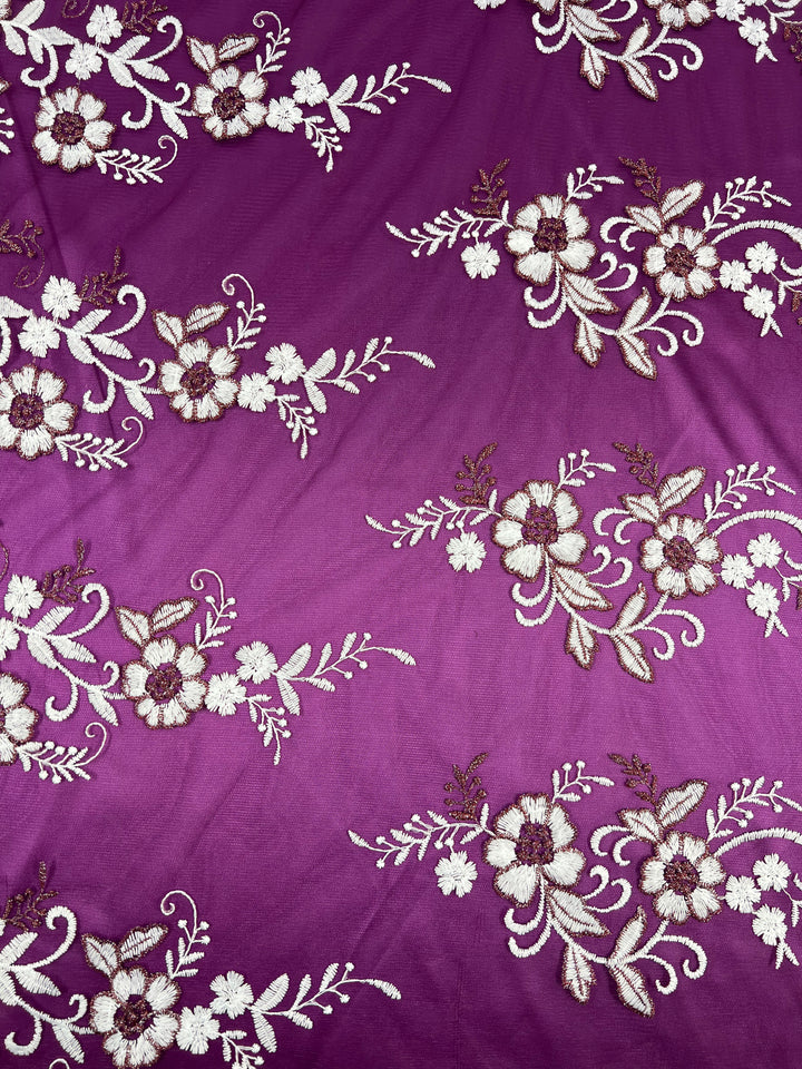 A purple fabric with embroidered floral patterns in white and beige. The intricate designs are evenly spaced, featuring flowers, leaves, and swirling vines. This Super Cheap Fabrics Embroidered Lace - Burgundy - 150cm lightweight fabric adds a delicate and elegant touch to the material, reminiscent of burgundy lace arrangements.