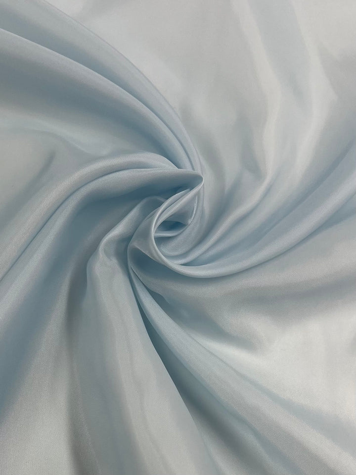 A close-up image of smooth, light blue satin fabric arranged in a swirl pattern. The vibrant fabric's glossy texture and delicate folds reflect light, giving a sense of softness and luxury, reminiscent of Super Cheap Fabrics' high-quality Lining - Sky Blue - 120cm.