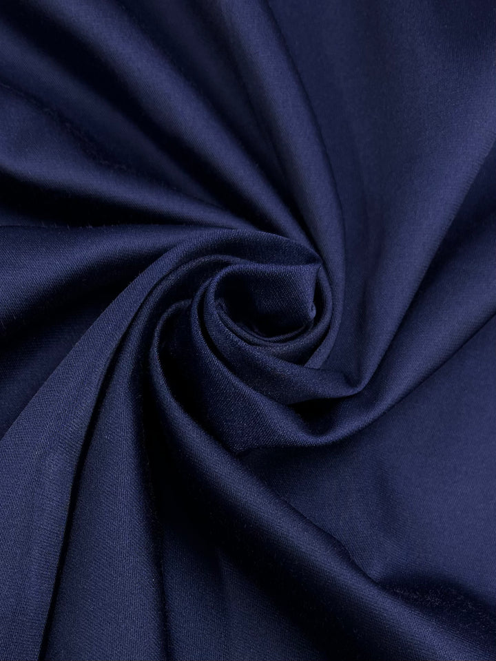 A close-up of Delustered Satin - Midnight Sail - 150cm by Super Cheap Fabrics with a smooth texture, arranged in an artistic swirl pattern at the center. The folds of the polyester fabric create gentle shadows and highlights, showcasing its soft and luxurious appearance, perfect for formal wear.