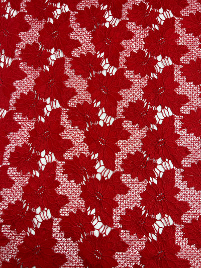 Close-up of a heavyweight fabric with a repeating pattern of red lace flowers, each with multiple petals. The flowers are interconnected by a delicate lace netting, creating an intricate and decorative design. The background is partially visible through the Super Cheap Fabrics Designer Anglaise Lace - Scarlet - 116cm polyester lace.