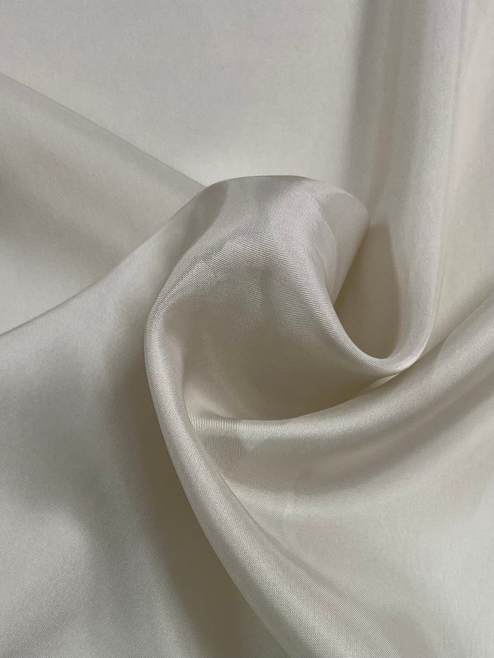 A close-up of a piece of smooth, lightweight fabric draped in gentle folds. The texture appears soft and slightly shiny, reflecting light subtly across its surface, reminiscent of materials used in elegant jackets or flowing skirts. This is the Lining - Natural - 120cm by Super Cheap Fabrics.