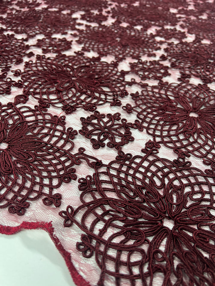 A close-up of intricate burgundy lace fabric with floral patterns embroidered on a light background. The Evening Lace - Elizabeth - by Super Cheap Fabrics features detailed, symmetrical designs and a textured surface, highlighting the delicate craftsmanship and ornamental nature of the fabric.