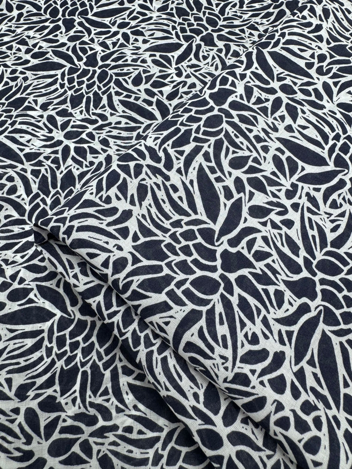 Close-up of Super Cheap Fabrics' "Cotton Voile - Lotus - 147cm," showcasing a black and white floral pattern. This lightweight, intricately patterned fabric is ideal for warm weather clothing, with densely packed flowers and leaves creating an abstract look that adds depth when folded.