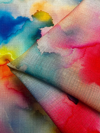 Close-up of the Super Cheap Fabrics' Designer Cotton - Venus - 150cm, ideal for luxury sewing projects. This fabric is artistically draped and features vibrant hues of red, blue, yellow, and green, showcasing the flowing and blending textures of watercolor paint.