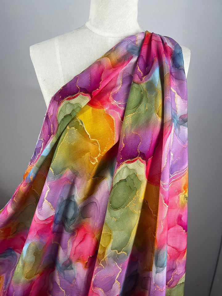 On display is a mannequin adorned with Designer Cotton - Adelina by Super Cheap Fabrics. This exquisite fabric exhibits a vivid array of pink, purple, yellow, blue, and green swirls in an abstract pattern, making it ideal for luxury sewing projects. Its sheer texture adds an artistic touch to any creation.