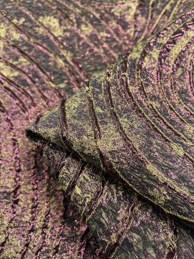 Close-up of the Designer Metallic Brocade - Amelia fabric from Super Cheap Fabrics, showcasing its lightweight texture with an organic, wave-like pattern. This fabric, sourced from Turkey, boasts a glittery, metallic finish in a mix of greens and purples, lending it a shimmering, iridescent appearance. The width of the fabric is 150cm.