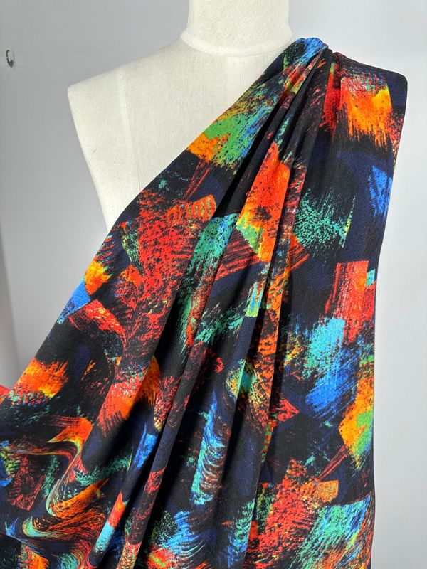 The Textured Knit - Art Attack by Super Cheap Fabrics is displayed on a mannequin, showcasing its vibrant, multicolored medium weight texture with abstract patterns in shades of red, green, blue, orange, and yellow against a black background. The fabric offers a smooth feel and generous stretch for a dynamic and artistic appearance.