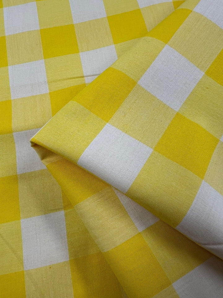 A close-up of the Cotton Gingham XL by Super Cheap Fabrics showcases a folded fabric featuring a yellow and white checkered pattern. Made from natural cotton fibers, this 112cm wide fabric displays evenly aligned squares, resulting in a bright, cheerful design that is soft and breathable.