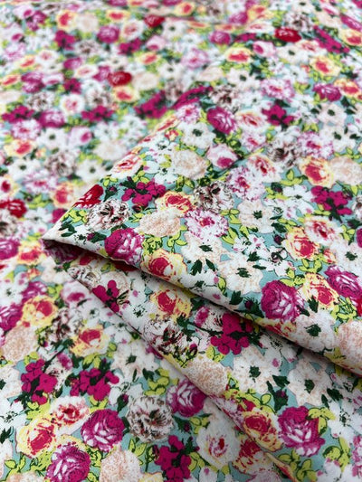 Close-up of Super Cheap Fabrics' "Printed Cotton - Roseness - 120cm," showcasing a vibrant pattern of red, pink, and yellow flowers with green leaves on a light background. The natural cotton fibers are slightly folded, highlighting the fabric's texture and colorful design ideal for various uses.