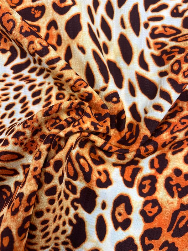 Close-up of the Deluxe Print - Jungle Tides - 155cm fabric by Super Cheap Fabrics, showcasing a polyester material with an animal print pattern reminiscent of leopard spots. The design highlights a blend of black and brown spots set against a background that shifts from light beige to warm orange tones. The fabric appears slightly wrinkled and draped, embodying the essence of versatile textiles.