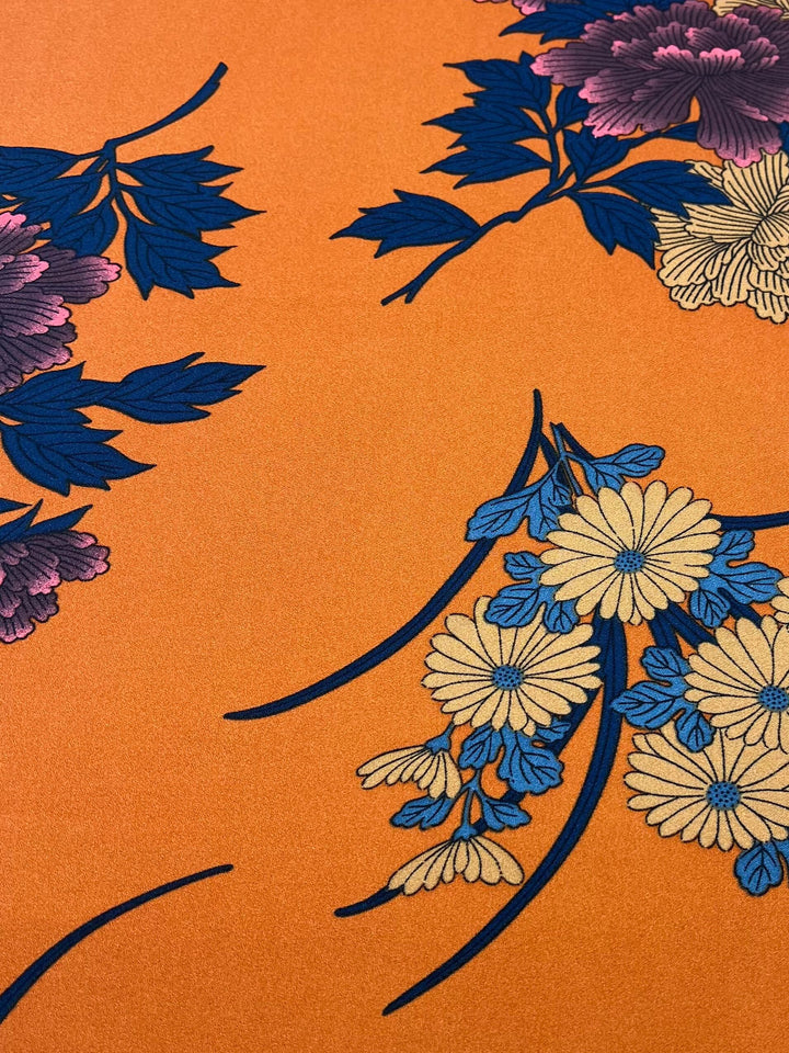 The Printed Crepe - Tuscan Flor by Super Cheap Fabrics is a 150cm wide orange polyester fabric with intricate floral patterns, featuring blue and pink leaves and clusters of yellow, blue, and pink flowers. Its elegant design is ideal for crafting high-fashion pieces.