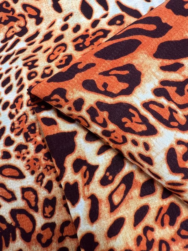 Close-up of folded Deluxe Print - Jungle Tides - 155cm polyester fabric from Super Cheap Fabrics, showcasing a vibrant leopard print pattern. The design features various shades of orange, brown, and black, creating an abstract and dynamic appearance. The texture and vivid colors give this versatile fabric a lifelike and striking look.