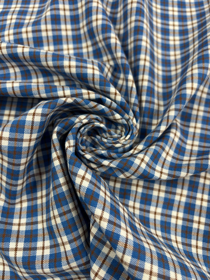 A detailed view of the Shirting - Ralph - 150cm fabric from Super Cheap Fabrics shows a swirl of blue, white, and brown plaid polyester. The lightweight design features intersecting lines that form squares, adding texture to its appearance. With its classic and rustic aesthetic, this versatile and soft fabric is ideal for various uses.