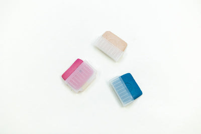 Three colorful Tailors Chalk in Holder - Pink by Super Cheap Fabrics with protective covers are arranged against a plain white background. The top chalk holder is beige, covered in transparent plastic, the bottom left chalk holder is pink, and the bottom right chalk holder is blue, both with white plastic partial covers.