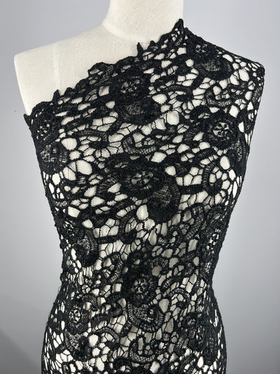 A mannequin displays a versatile one-shoulder garment made of intricate Anglaise Lace - Black Garden - 115cm from Super Cheap Fabrics with floral patterns. The design features semi-sheer areas with detailed, intertwining motifs, creating an elegant and sophisticated look against a light background.