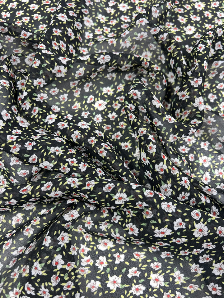 The "Super Cheap Fabrics" Printed Georgette - Dark Blooming Seeds - 150cm features a close-up of black Georgette fabric adorned with a small floral print showcasing painted flowers in white, red, and yellow hues, complemented by green leaves. The sheer polyester material appears slightly crumpled, exhibiting light variations in texture and wrinkles.