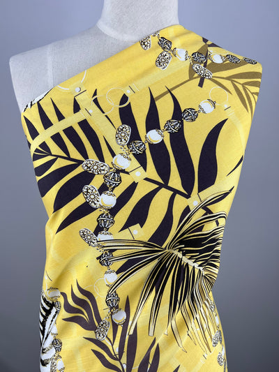 A vibrant yellow, lightweight fabric draped over a mannequin, featuring a bold black and white tropical leaf and floral pattern accented with geometric shapes. The design is asymmetrical, wrapping around the dummy with a single shoulder exposed. The fabric used is Bamboo Rayon - Pina Colada - 150cm from Super Cheap Fabrics.