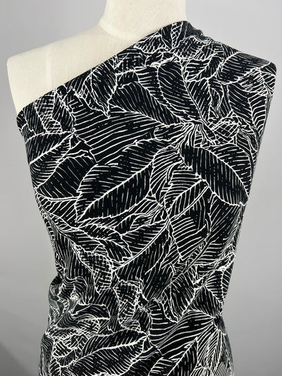 A dress form draped in Deluxe Print - Leafy Layers - 150cm from Super Cheap Fabrics. The detailed, intricate leaves with fine lines create a striking contrast against the dark background. This versatile fabric is styled asymmetrically over the shoulder.