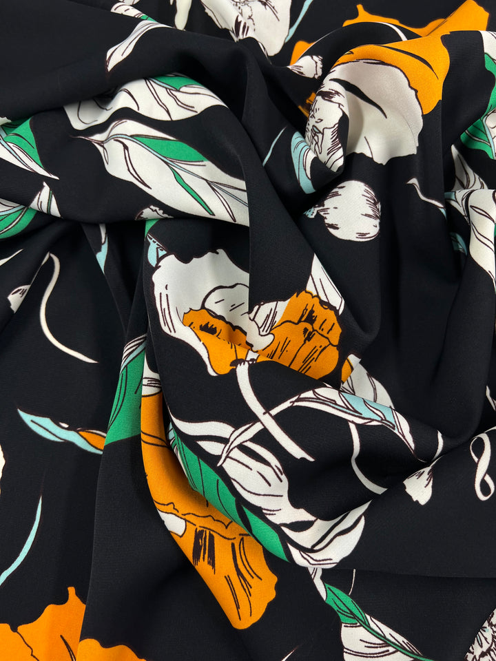 A close-up of a Deluxe Print - Marigold Comic - 150cm fabric by Super Cheap Fabrics with a floral pattern featuring orange, white, and green flowers and leaves. The fabric is gently folded, showcasing the intricate design and vibrant colors typical of designer fabrics.