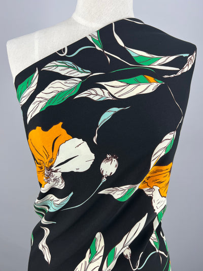 A garment on a mannequin featuring a black background adorned with a vibrant floral pattern crafted from versatile polyester fabrics. The design includes large, colorful flowers in shades of orange, green, and white with detailed leaves and stems, creating a striking and elegant look made from Deluxe Print - Marigold Comic - 150cm by Super Cheap Fabrics.