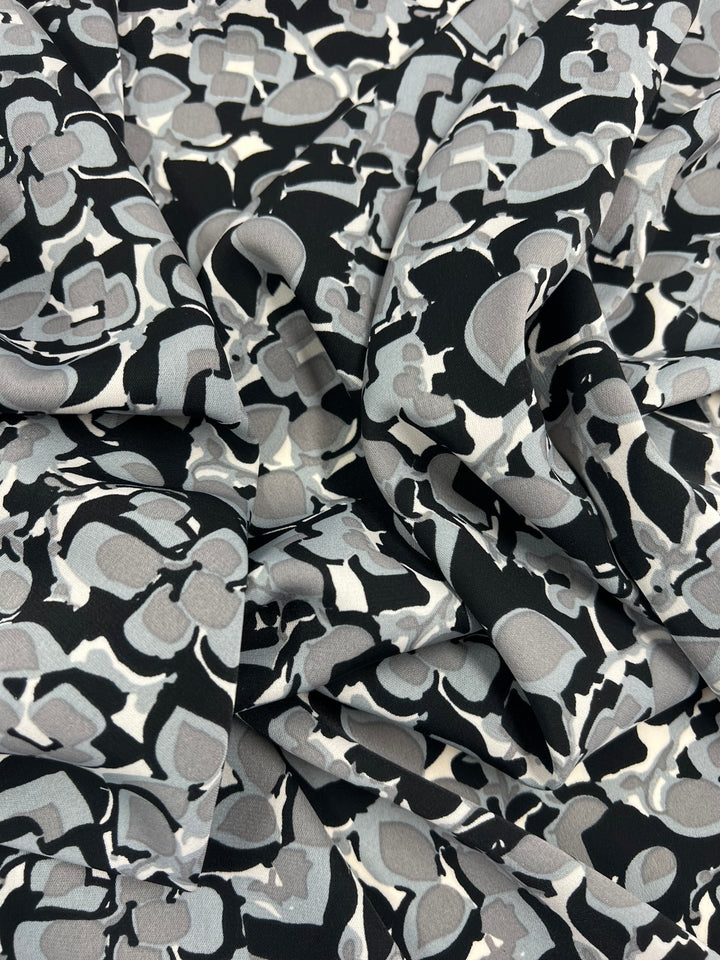 A close-up view of a crumpled fabric featuring a black base with an abstract pattern in shades of gray and white. The design consists of various shapes and forms, creating a visually dynamic and textured appearance, characteristic of Deluxe Print - Flower Blocks - 150cm by Super Cheap Fabrics often seen in designer collections.