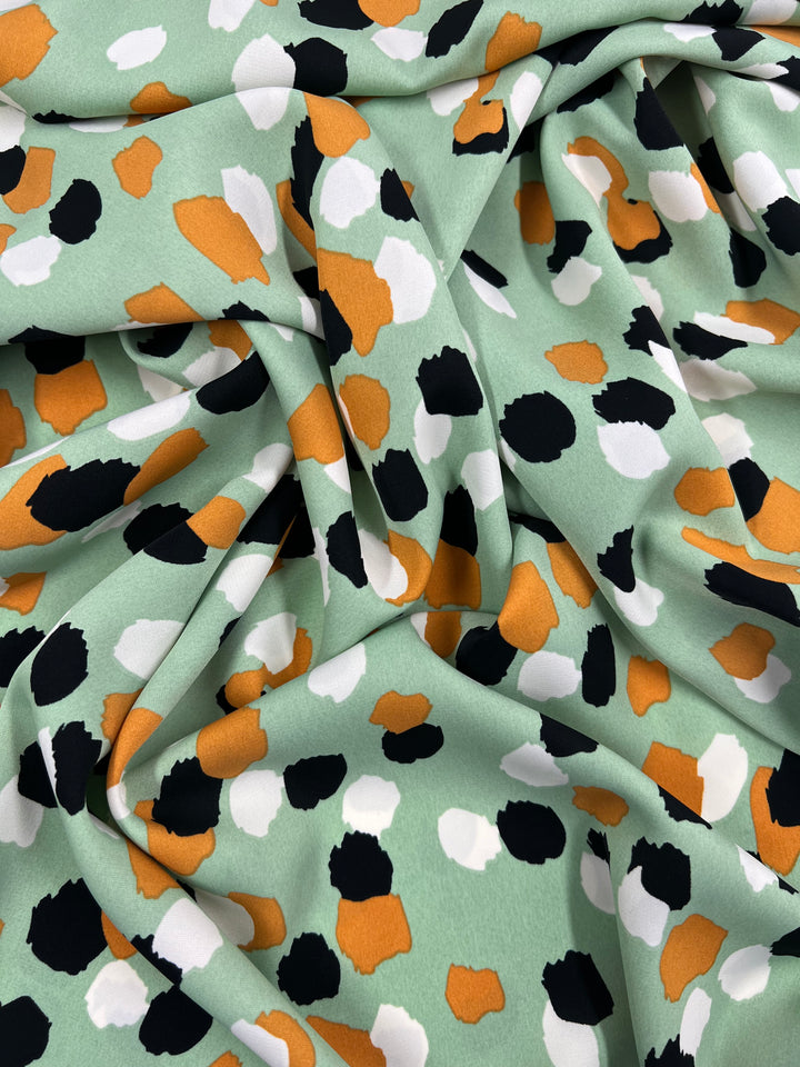 A soft polyester fabric featuring a colorful abstract pattern. The Super Cheap Fabrics Deluxe Print - Painters Palette - 150cm has a mint green background with irregular spots in orange, black, and white, creating a lively and dynamic design. Perfect for versatile fabrics, it is arranged in gentle folds.