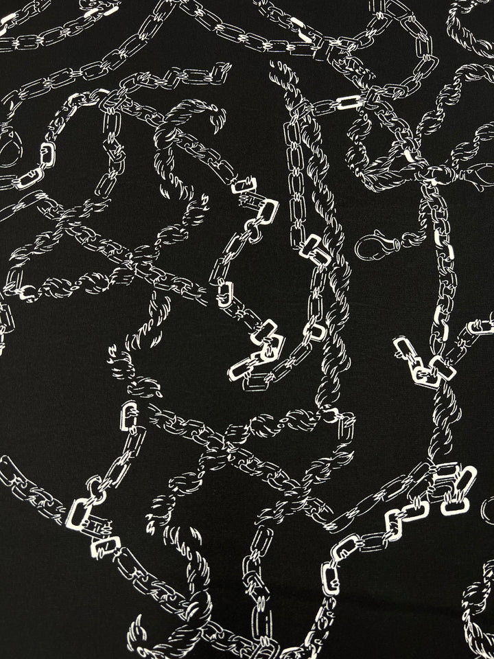 An illustration of white chains intertwined and overlapping on a solid black background, resembling the intricate patterns often found in designer fabrics. Each chain is detailed with small links, creating a chaotic yet meticulously crafted design that evokes the complexity of Deluxe Print - MonoLinks - 150cm by Super Cheap Fabrics.